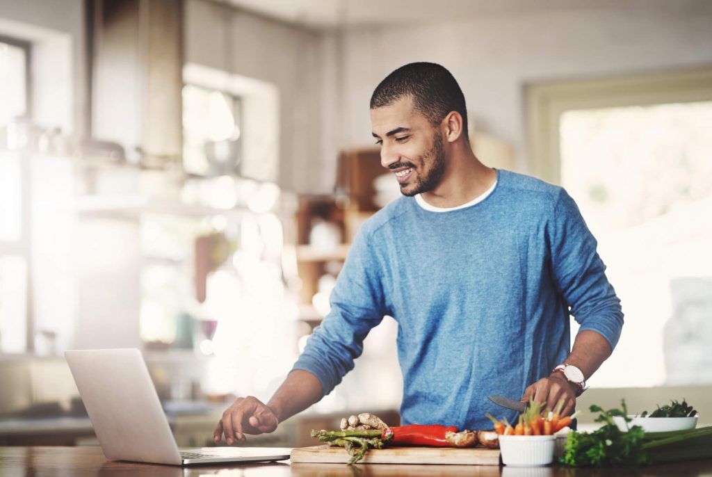 A man on his laptop in the kitchen chopping vegetables